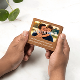 A person holds a small brown leather photo card holder with a customizable sample photo of a father and son and the printed quote, 'Dad, wherever life takes you, a little piece of me goes with you.' on a white marble surface with green leaves in the background.