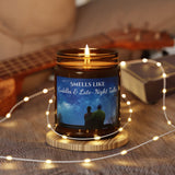 Candle in a jar with a label featuring two people and text, surrounded by string lights and a guitar in the background.