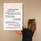 A woman in a black shirt holds a large personalized wall art print against a beige wall. The print has a white background with a faint pink crown at the top and a printed poem titled 'My Little PRINCESS' addressed to a child's name.