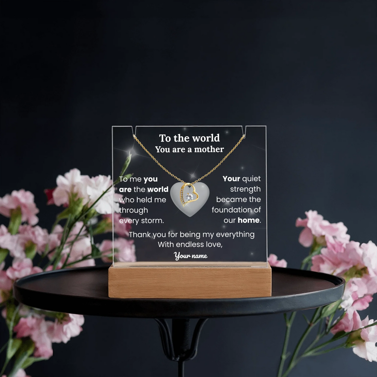 Acrylic plaque with necklace on a stand, surrounded by flowers against a dark background