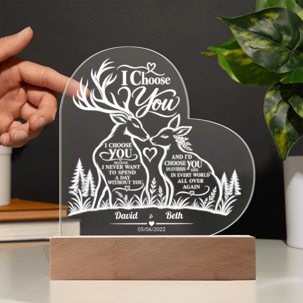 Personalized acrylic laser-engraved wedding plaque featuring deer and forest motifs with the text 'I Choose You' and custom names 'David & Beth' on a wooden base. A hand is holding the left side of the plaque, with a plant and shelf in the background.