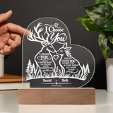 Personalized acrylic laser-engraved wedding plaque featuring deer and forest motifs with the text 'I Choose You' and custom names 'David & Beth' on a wooden base. A hand is holding the left side of the plaque, with a plant and shelf in the background.