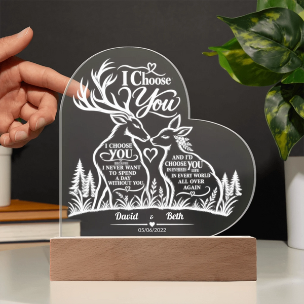 Personalized acrylic laser-engraved wedding plaque featuring deer and forest motifs with the text 'I Choose You' and custom names 'David & Beth' on a wooden base. A hand is holding the left side of the plaque, with a plant and shelf in the background.