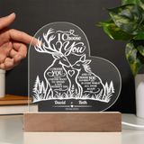Personalized acrylic LED night light with deer and forest design, held by a hand on a wooden base with a dark background and green plant leaves.