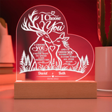 Personalized acrylic LED night light shaped like a heart, featuring two deer facing each other with romantic engraved text and names 'David & Beth' on a wooden base, shown glowing in red on a tabletop.