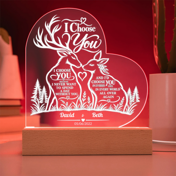 Personalized acrylic LED night light shaped like a heart, featuring two deer facing each other with romantic engraved text and names 'David & Beth' on a wooden base, shown glowing in red on a tabletop.