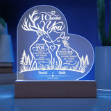 Personalized 3D LED acrylic night light shaped like a heart, featuring two deer and romantic text, glowing on a wooden base in a blue-lit room.