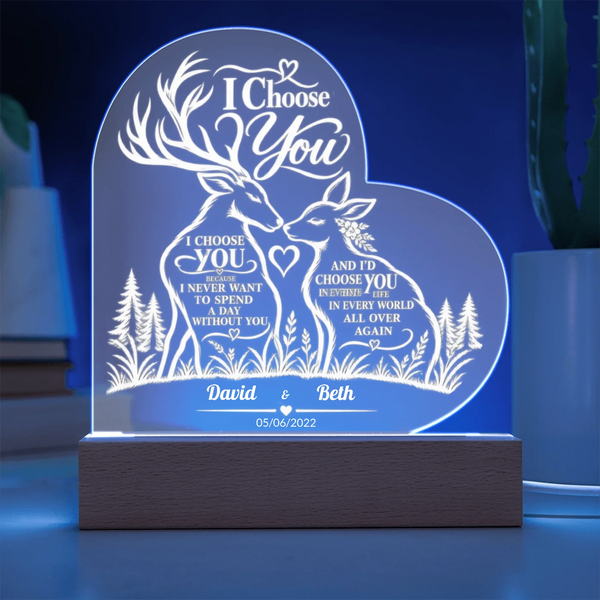 Personalized 3D LED acrylic night light shaped like a heart, featuring two deer and romantic text, glowing on a wooden base in a blue-lit room.