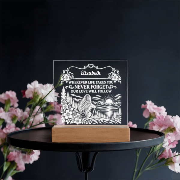 A personalized acrylic keepsake plaque with an engraved silhouette of a woman and flowers, resting on a light wood block base. The plaque is displayed on a black round table, framed by pink flowers against a dark background.