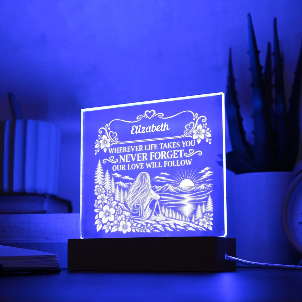 A personalized acrylic 3D LED night light glowing with a blue light, featuring an engraved illustration of a woman by a lake and the name 'Elizabeth' with a sentimental message. It sits on a dark wooden base on a table with a blurred background of books and a plant.