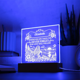 A personalized acrylic 3D LED night light glowing with a blue light, featuring an engraved illustration of a woman by a lake and the name 'Elizabeth' with a sentimental message. It sits on a dark wooden base on a table with a blurred background of books and a plant.