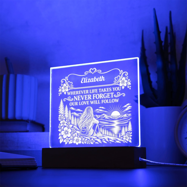 A personalized acrylic 3D LED night light glowing with a blue light, featuring an engraved illustration of a woman by a lake and the name 'Elizabeth' with a sentimental message. It sits on a dark wooden base on a table with a blurred background of books and a plant.