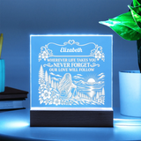 A personalized acrylic LED night light on a dark wooden base, glowing with a blue light. The light features an engraved design of a woman camping by a lake with mountains and flowers, with the name 'Elizabeth' and a memorial quote.