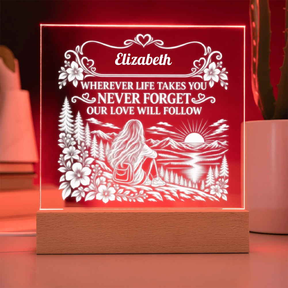 A personalized acrylic LED night light on a wooden base, glowing with a warm red light. The front features a white laser-cut design of a woman sitting in a forest by a lake at sunset, with the name 'Elizabeth' and the message 'WHEREVER LIFE TAKES YOU NEVER FORGET OUR LOVE WILL FOLLOW' at the top.