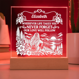 A personalized acrylic LED night light on a wooden base, glowing with a warm red light. The front features a white laser-cut design of a woman sitting in a forest by a lake at sunset, with the name 'Elizabeth' and the message 'WHEREVER LIFE TAKES YOU NEVER FORGET OUR LOVE WILL FOLLOW' at the top.