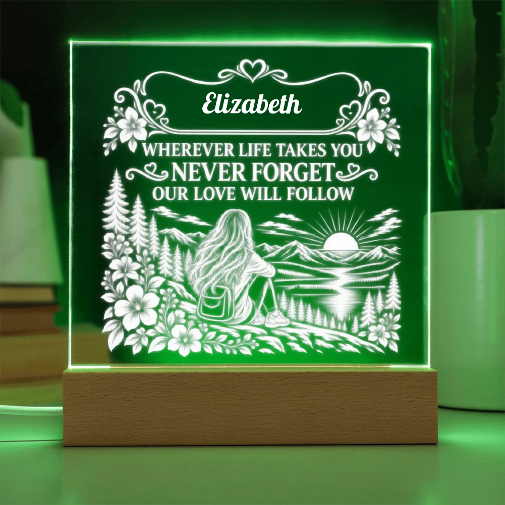 A personalized LED acrylic night light on a wooden base, glowing with a green light. The light features a white laser-engraved design of a woman sitting by a lake with mountains and a sunset, with the name 'Elizabeth' and a sentimental message above the scene.