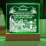 A personalized LED acrylic night light on a wooden base, glowing with a green light. The light features a white laser-engraved design of a woman sitting by a lake with mountains and a sunset, with the name 'Elizabeth' and a sentimental message above the scene.