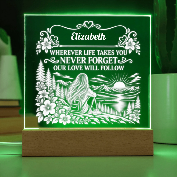 A personalized LED acrylic night light on a wooden base, glowing with a green light. The light features a white laser-engraved design of a woman sitting by a lake with mountains and a sunset, with the name 'Elizabeth' and a sentimental message above the scene.