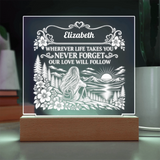A personalized acrylic LED night light on a wooden base, featuring a white engraved design of a woman sitting by a lake with mountains and flowers, with the text 'Elizabeth WHEREVER LIFE TAKES YOU NEVER FORGET OUR LOVE WILL FOLLOW'.