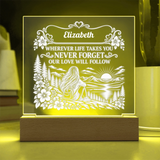 Personalized acrylic LED night light with a wooden base, featuring a scenic engraving of a woman sitting by a lake with mountains and flowers, and the text 'Elizabeth WHEREVER LIFE TAKES YOU NEVER FORGET OUR LOVE WILL FOLLOW', glowing with a warm yellow light.