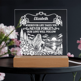 Personalized acrylic LED night light with a wooden base, featuring a engraved illustration of a woman sitting by a lake with the text 'Elizabeth WHEREVER LIFE TAKES YOU NEVER FORGET OUR LOVE WILL FOLLOW', displayed on a dark table with blurred pink flowers.