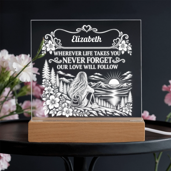 Personalized acrylic LED night light with a wooden base, featuring a engraved illustration of a woman sitting by a lake with the text 'Elizabeth WHEREVER LIFE TAKES YOU NEVER FORGET OUR LOVE WILL FOLLOW', displayed on a dark table with blurred pink flowers.