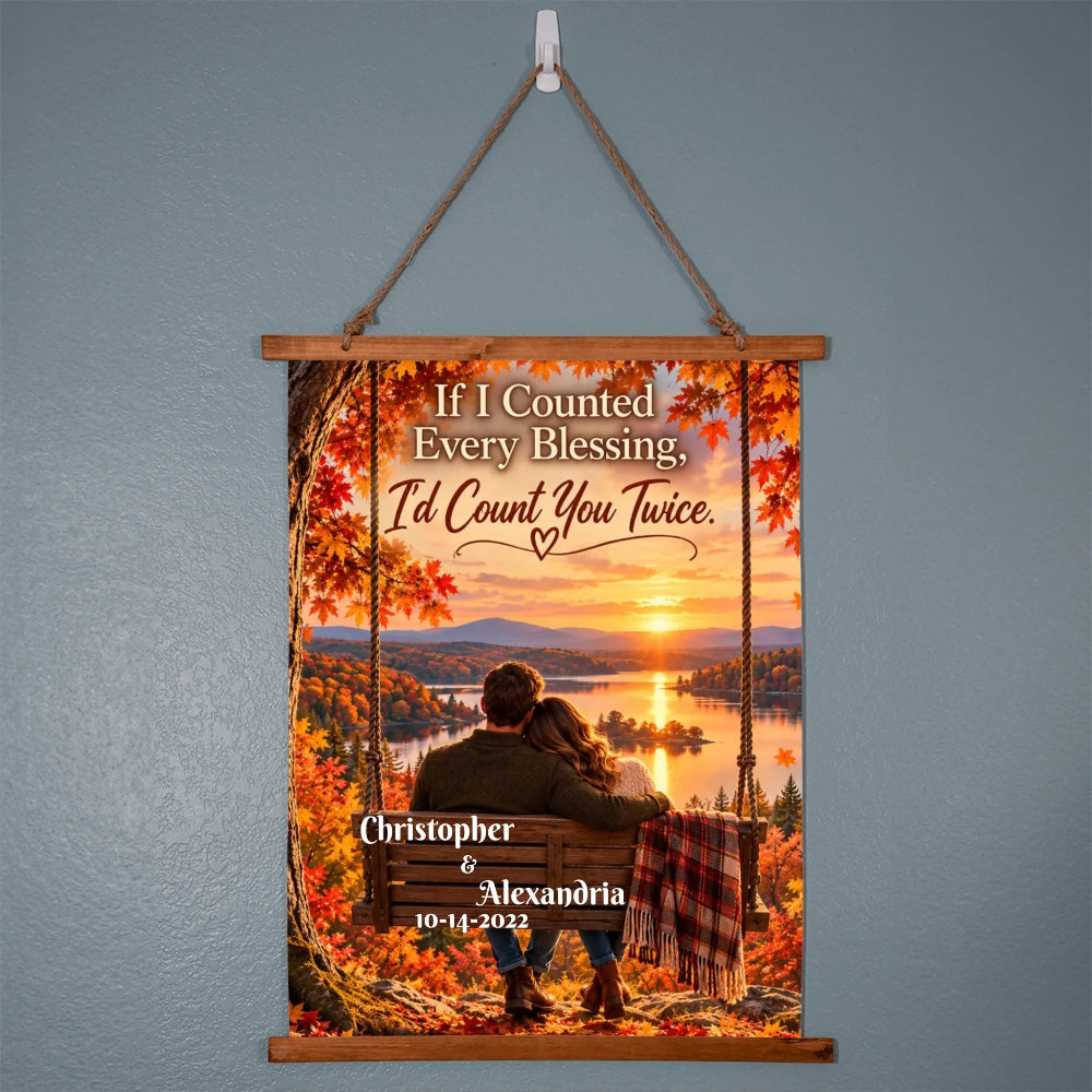 A personalized hanging wall scroll poster with a wooden top and bottom frame, suspended by a rope from a white wall hook. The scroll features a scenic autumn sunset landscape with a couple sitting on a bench by the water, surrounded by colorful fall leaves.