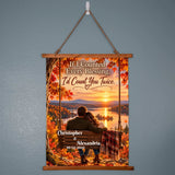 A personalized hanging wall scroll poster with a wooden top and bottom frame, suspended by a rope from a white wall hook. The scroll features a scenic autumn sunset landscape with a couple sitting on a bench by the water, surrounded by colorful fall leaves.