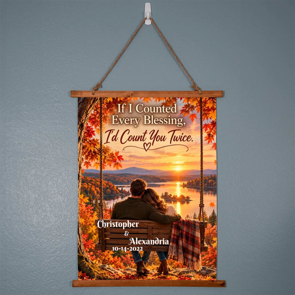 A personalized hanging wall scroll poster with a wooden top and bottom frame, suspended by a rope from a white wall hook. The scroll features a scenic autumn sunset landscape with a couple sitting on a bench by the water, surrounded by colorful fall leaves.