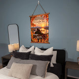 A vertical wall hanging poster with a wooden top frame and rope hanger, displayed above a bed in a styled bedroom. The poster features an autumn landscape with red and orange leaves, a sunset over water, and a couple sitting by the lake.