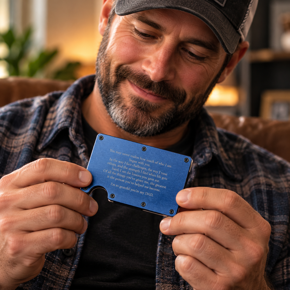 A man in a plaid shirt and baseball cap holds a small blue metal card with engraved text in both hands, looking down at it in a warmly lit indoor setting.