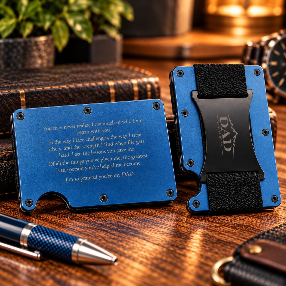 A blue metal card holder wallet shown from two angles on a wooden desk, with engraved text on the front and a black clip holding a card. A pen and leather accessories are visible in the background.