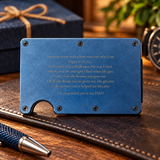 A blue metal slim wallet card holder engraved with a heartfelt message for a DAD, resting on a brown leather surface with a pen in the foreground and a blurred gift box in the background.