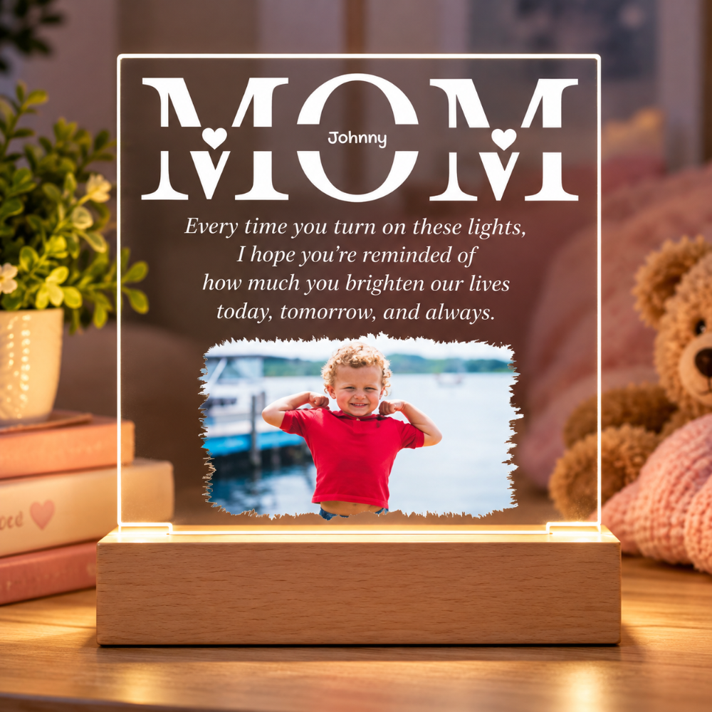 A personalized acrylic light plaque on a wooden base, illuminated from within. The plaque features the word 'MOM' with 'Johnny' in the center and a heart detail, above a printed quote.