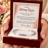 A silver open cuff bracelet presented in a dark red wooden gift box with a white insert card that reads 'To The Woman I Love' and includes a placeholder text area for the bracelet. Hands with pink manicured nails hold the box.