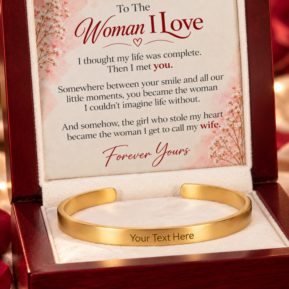 A gold cuff bracelet displayed inside an open wooden gift box with a printed message card. The card reads 'To The Woman I Love' and includes a sentimental message, while the bracelet has 'Your Text Here' engraved on the inside.