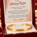 A gold cuff bracelet displayed inside an open wooden gift box with a printed message card. The card reads 'To The Woman I Love' and includes a sentimental message, while the bracelet has 'Your Text Here' engraved on the inside.
