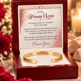 An open wooden gift box with a red interior lid displaying a sentimental message card and a gold open cuff bracelet on a white cushion. The box is photographed in a warm, softly blurred setting with flowers and candles in the background.