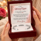 A silver open cuff bracelet displayed inside a dark red wooden gift box with a sentimental message card that reads 'To The Woman I Love'. The box is held by two hands against a warm, blurred background with a pink rose.
