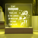A wooden LED acrylic night light with a glowing yellow-green ambiance, featuring a white laser-engraved illustration of a couple hugging and the text 'TO MY HUSBAND YOUR LOVE IS THE CANVAS ON WHICH MY DREAMS COME ALIVE.'.