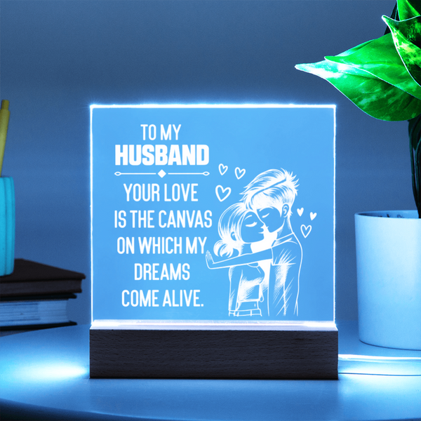 A glowing acrylic LED sign on a dark wooden base, featuring white text that reads 'TO MY HUSBAND YOUR LOVE IS THE CANVAS ON WHICH MY DREAMS COME ALIVE' with a couple illustration and small hearts, displayed on a tabletop with ambient blue lighting.