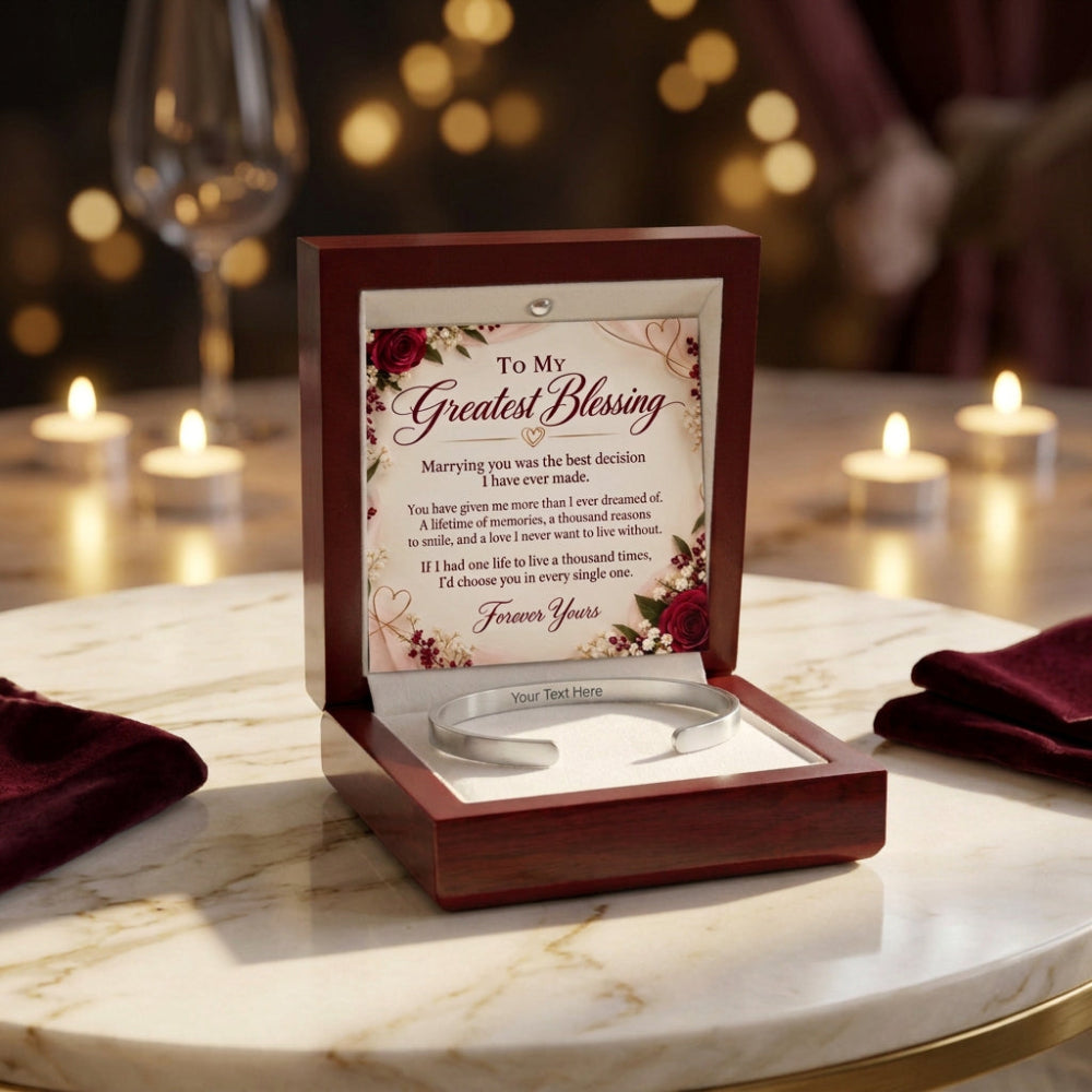 An open wooden keepsake box on a marble table, displaying a silver band with a custom engraving beneath a printed message card that reads 'To My Greatest Blessing.' The scene is styled with lit candles, blurred wine glasses, and burgundy napkins in the background.