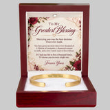 A personalized gold cuff bracelet presented in a dark reddish-brown wooden gift box with a floral message card inside. The card reads 'To My Greatest Blessing' and includes a gold bracelet with placeholder text 'Your Text Here' on a white background.