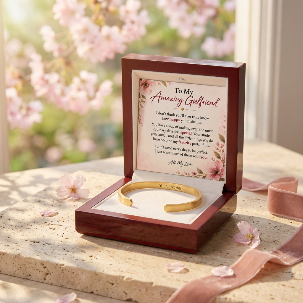 An open dark wood gift box on a stone surface with cherry blossoms and pink petals in the background. Inside is a gold bracelet on a white insert with a printed message card reading 'To My Amazing Girlfriend.