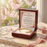 An open dark wood gift box on a stone surface with cherry blossoms and pink petals in the background. Inside is a gold bracelet on a white insert with a printed message card reading 'To My Amazing Girlfriend.
