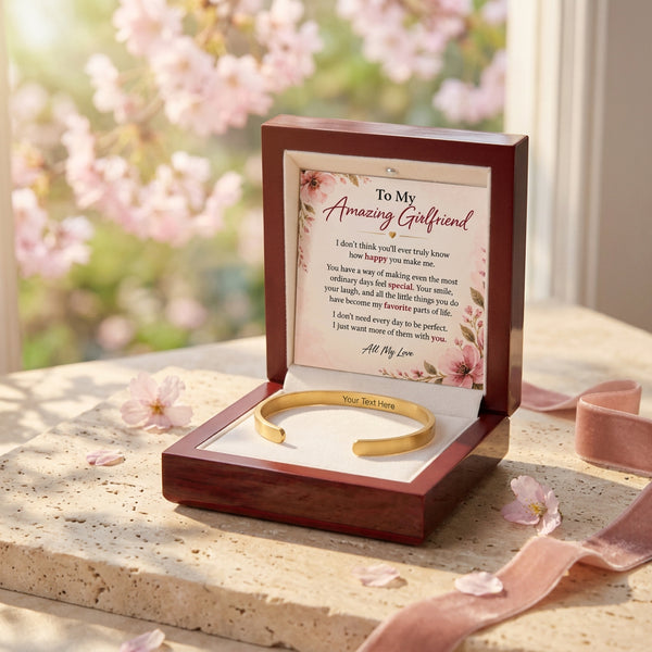 An open dark wood gift box on a stone surface with cherry blossoms and pink petals in the background. Inside is a gold bracelet on a white insert with a printed message card reading 'To My Amazing Girlfriend.