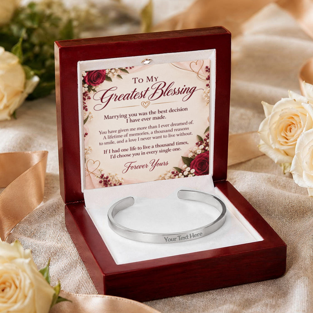 A silver engraved cuff bracelet presented in an open dark red wooden gift box with a floral message card inside. The box sits on a beige fabric surface with cream roses in the background.