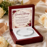 A silver engraved cuff bracelet presented in an open dark red wooden gift box with a floral message card inside. The box sits on a beige fabric surface with cream roses in the background.