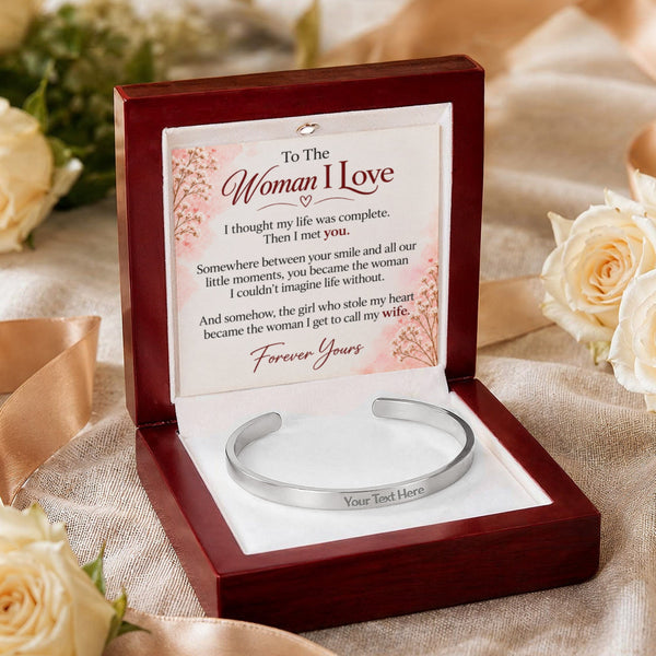 A silver engraved cuff bracelet presented in an open dark wood gift box with a printed message card inside, styled with cream roses on a beige fabric background.