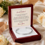 A silver open cuff bracelet displayed inside a dark red wooden gift box with a printed message card. The box sits on a beige fabric surface with soft cream roses in the background.